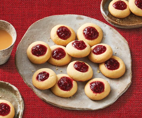 Overhead image of home-baked raspberry thumbprint cookies, on three different sized plates with a complementary hot beverage on the left side.