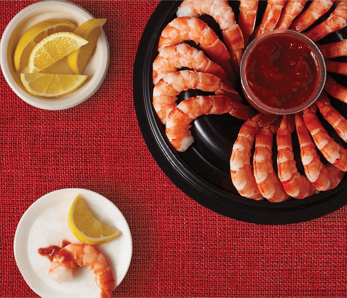 Image of a pack shot of an open Compliments Shrimp Ring with Cocktail Sauce, a plate of lemon wedges and a small plate with a single shrimp and slice of lemon on top.
