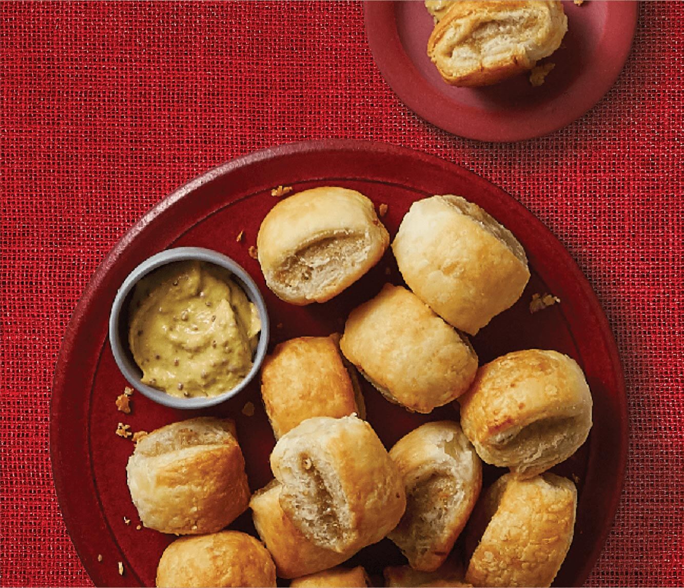 A red large red plate of golden baked sausage rolls with a dipping sauce, and a smaller red plate withn a single sausage roll in the upper right hand corner.
