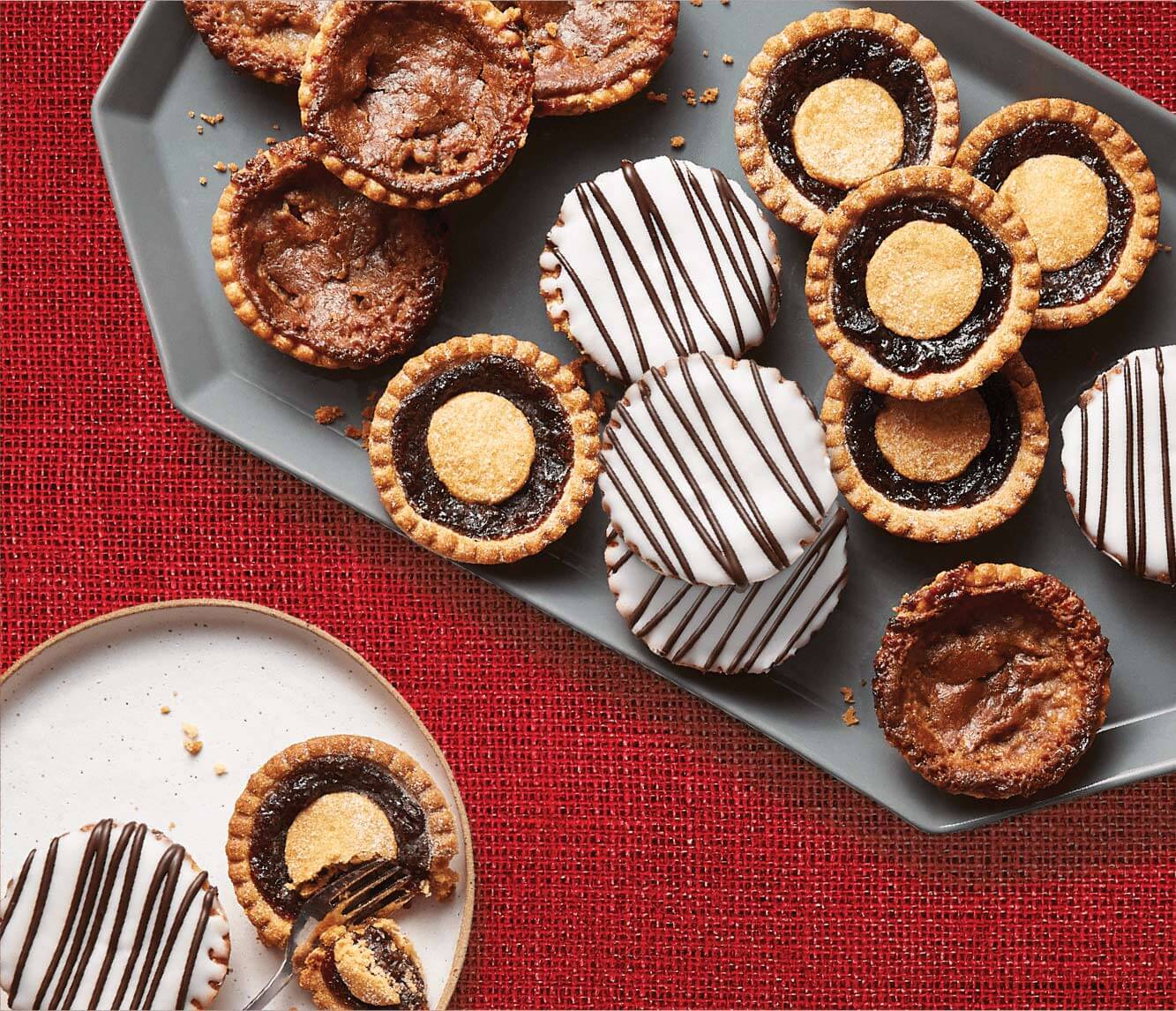 One grey serving platter with a stack of butter tarts in various flavours and a smaller white plate with two butter tarts in the left hand bottom corner. 