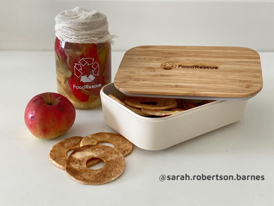 A reusable container on a table filled with apple chips, a fresh apple beside it and a jar of apples behind.