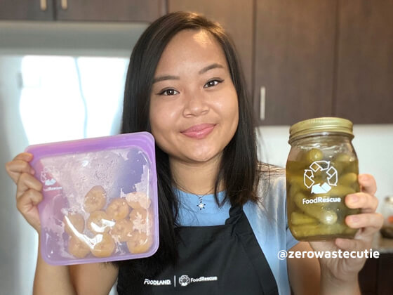 Elizabeth stands in her kitchen in an apron and shows off a jar of pickles and a reusable bag with snacks inside. Fresh fruits are in front of her.
